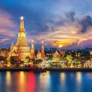 Un complexe de temples orné et illuminé, doté de multiples flèches, se dresse au bord d'une rivière au coucher du soleil, ses lumières se reflétant sur l'eau.