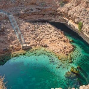 Una lunga scalinata scende in una conca rocciosa piena di acqua cristallina color turchese, incastonata tra scogliere aride.