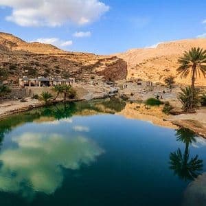 Un'oasi nel deserto con acqua calma e blu che riflette palme e un canyon roccioso sotto un cielo parzialmente nuvoloso.