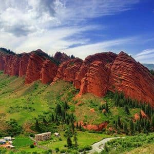Formazioni rocciose rosse frastagliate dominano una lussureggiante valle verde con un piccolo villaggio e un fiume sotto un cielo parzialmente nuvoloso.