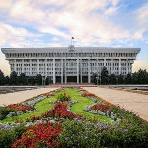 A large, symmetrical government building with a flag, seen from across a plaza with colorful, manicured flowerbeds.