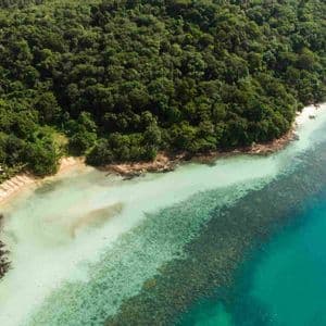 Vista aerea di un'isola tropicale lussureggiante con piccole case adagiate su una spiaggia sabbiosa e acqua turchese cristallina.