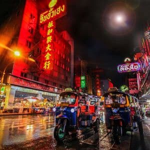 Due tuk-tuk sono parcheggiati di notte su una strada cittadina bagnata, illuminati dal bagliore intenso di insegne al neon rosse e gialle.