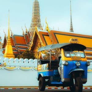 Un tuk-tuk blu e giallo parcheggiato su una strada davanti a un tempio decorato con guglie dorate e tetti arancioni.