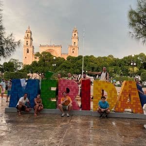 Un viaje en grupo de WeRoad posando para una foto con las grandes y coloridas letras que forman MÉRIDA en una plaza con una catedral al fondo.