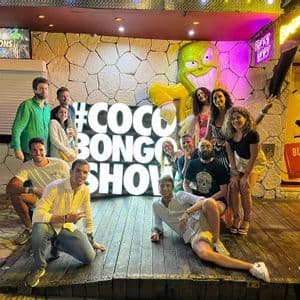 A WeRoad group trip poses for a photo at night in front of a large, illuminated sign that reads '#COCO BONGO SHOW'.