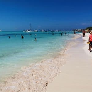 Many people enjoy a sunny day at a beach with white sand and clear turquoise water, with several boats on the horizon.