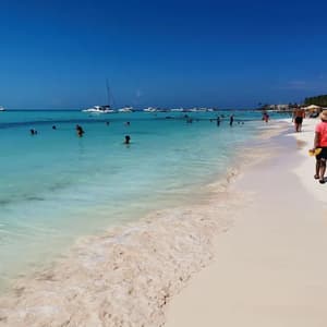 Un día soleado en una playa concurrida donde la gente nada en agua turquesa y camina sobre arena blanca, con barcos visibles en la distancia.