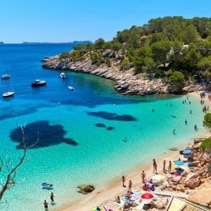 An aerial view of a busy beach cove with people swimming in turquoise water and boats anchored in the bay under a clear blue sky.