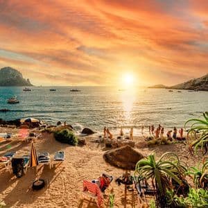 Un atardecer naranja vibrante sobre una cala de playa llena de gente, con barcos en el agua y una gran isla rocosa en la distancia.
