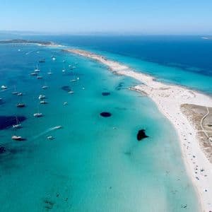 An aerial view of numerous sailboats anchored in clear turquoise waters alongside a long white sand spit.