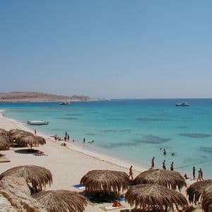 Blick von oben auf einen tropischen Strand mit Strohschirmen, Badenden im türkisfarbenen Wasser und Booten in der Ferne.