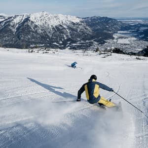 Ein Skifahrer carvt von hinten einen verschneiten Hang hinunter, wirbelt Schnee auf, mit einem weiten Gebirgstal im Hintergrund.