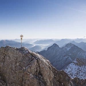 Ein goldenes Gipfelkreuz auf einem felsigen Gipfel überblickt eine weite Bergkette unter strahlender Sonne und klarem Himmel.
