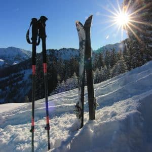 Ein Paar Ski und Skistöcke stecken aufrecht im Tiefschnee auf einem sonnigen Berg, mit einem hellen Sonnenstern am Bildrand.
