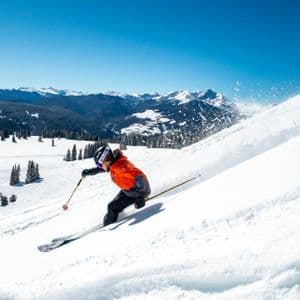 Ein Skifahrer in orangefarbener Jacke carvt einen Pulverschneehang hinunter, wirbelt dabei Schnee auf, unter klarem blauem Himmel und mit fernen Bergen.