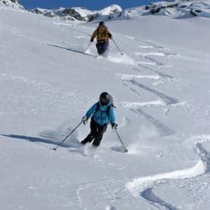 Zwei Personen einer WeRoad Gruppenreise skifahren einen sonnigen, schneebedeckten Berg hinunter und hinterlassen geschwungene Spuren im Pulverschnee.