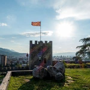 Die Flagge Nordmazedoniens weht von einem steinernen Burgturm auf einem Hügel, mit Blick auf eine Stadt und Berge unter strahlender Sonne.