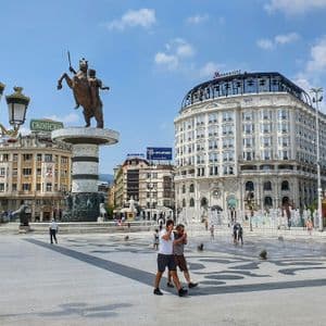 Eine große Reiterstatue steht auf einem Stadtplatz mit Springbrunnen, umgeben von Gebäuden und Menschen unter blauem Himmel.