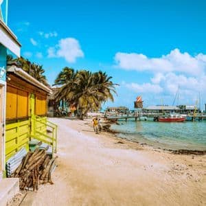 Bunte Holzgebäude und Palmen säumen einen Sandstrand neben einem Pier mit Booten auf türkisfarbenem Wasser.