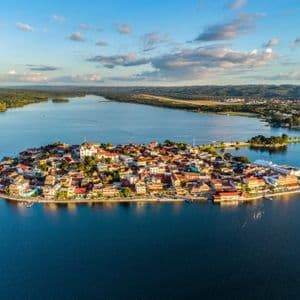 Eine Luftaufnahme einer kleinen, dicht besiedelten Inselstadt an einem blauen See, mit einem bewaldeten Festland im Hintergrund.
