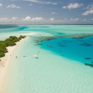 Una vista aerea di un banco di sabbia tropicale con vegetazione lussureggiante, circondato da acqua turchese dove le persone nuotano e le barche sono ancorate.