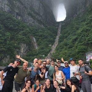 Un gruppo WeRoad in viaggio si fa fotografare ai piedi di una montagna ripida e verde, con un grande arco naturale e una lunga scalinata.