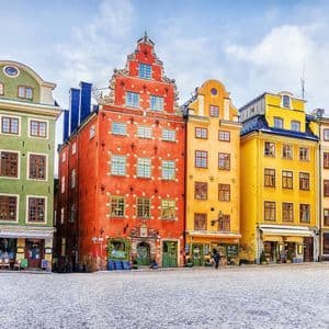 Una fila di colorati edifici storici a capanna si affacciano su una piazza acciottolata sotto un cielo azzurro e nuvoloso.