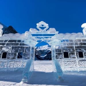 Un castello fatto di grandi blocchi di ghiaccio con un arco centrale si erge nella neve, con le montagne visibili sullo sfondo contro un cielo blu.
