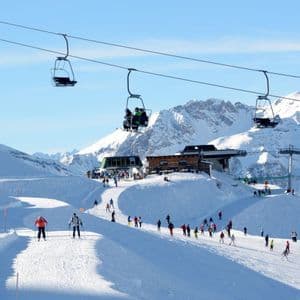 Sciatori su una pista affollata in una località montana, con una seggiovia che passa sopra sotto un cielo azzurro.