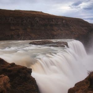 Una fotografia a lunga esposizione di una vasta cascata che precipita in un canyon nebbioso, fiancheggiato da scogliere rocciose marroni sotto un cielo nuvoloso.