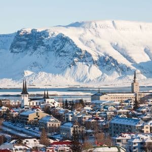 Una veduta aerea di una città innevata con tetti colorati e campanili di chiese, adagiata di fronte a un'imponente montagna innevata oltre uno specchio d'acqua.