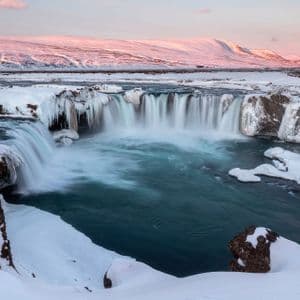 Una vasta cascata con i bordi ghiacciati si riversa in una piscina turchese in un paesaggio vasto e innevato al tramonto.