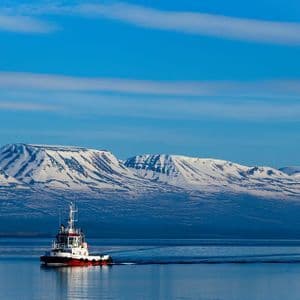Un rimorchiatore rosso e bianco naviga su acque calme e blu, con un'ampia catena montuosa innevata sullo sfondo sotto un cielo sereno.