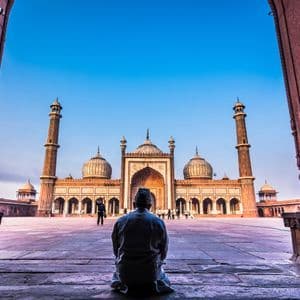 Un uomo visto di spalle si inginocchia all'interno di un arco di pietra, guardando verso una grande moschea con minareti al di là di un cortile.