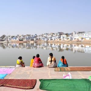 Cinco mujeres con saris coloridos se sientan en escalones de piedra junto a un lago, con textiles secándose en primer plano y una ciudad reflejada en el agua.