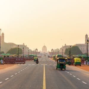 Des tuk-tuks et une moto circulent sur une large route à plusieurs voies, en direction de grands édifices à dôme, sous un ciel doré et brumeux.