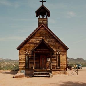 Una chiesa rustica in legno con campanile si erge in un paesaggio desertico sotto un cielo limpido.