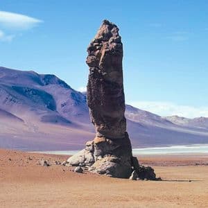Una guglia di roccia imponente si erge in un vasto deserto, con montagne viola in lontananza e una distesa salina sotto un cielo azzurro e limpido.