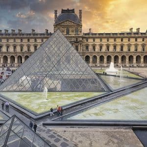 Una grande piramide di vetro e fontane circondate da persone in un cortile storico sotto un cielo caldo al tramonto.