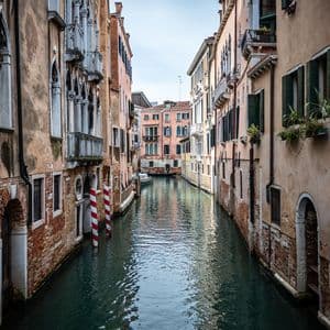 A narrow canal flows between tall, weathered buildings with balconies and shutters under an overcast sky.