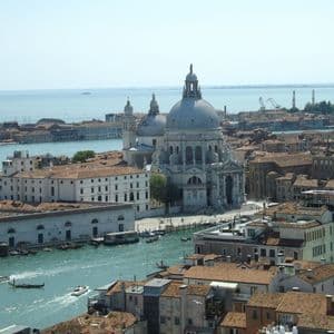 An aerial view of a city with a large domed basilica and terracotta-roofed buildings next to a canal with boats.