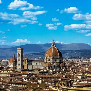 The Florence Cathedral's dome and bell tower rise above the city's terracotta roofs, with mountains in the background under a blue sky.