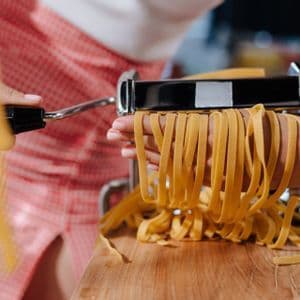 A person's hands catch strands of fresh fettuccine pasta coming out of a manual pasta maker over a wooden board.