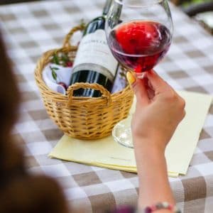 A person holds a glass of red wine over a checkered tablecloth with a wine bottle in a wicker basket.