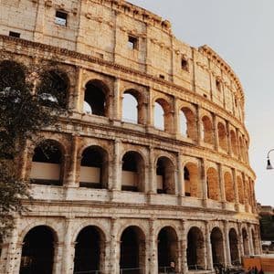The exterior of a large ancient stone amphitheater with multiple tiers of arches, illuminated by the setting sun.