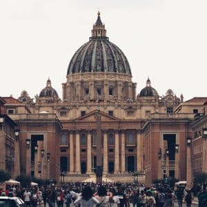 A large crowd gathers on a wide street leading to the grand facade and dome of a historic basilica under an overcast sky.