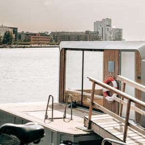 A modern floating wooden sauna with large glass windows and a lifebuoy rests on the water near an urban shoreline.