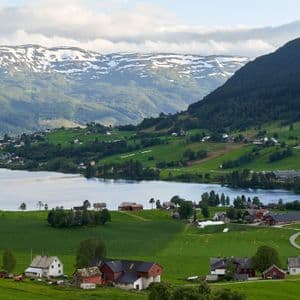 Rural houses and green farm fields along a lake with snow-capped mountains in the background.