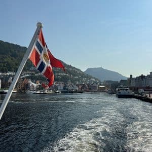 A boat leaves a waterfront harbor lined with historic buildings and green hills under a clear blue sky.